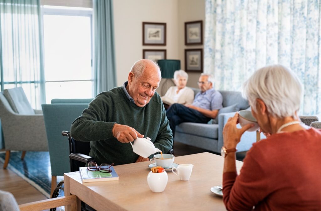 An older man in a wheelchair pouring tea at a table while socializing with a woman.