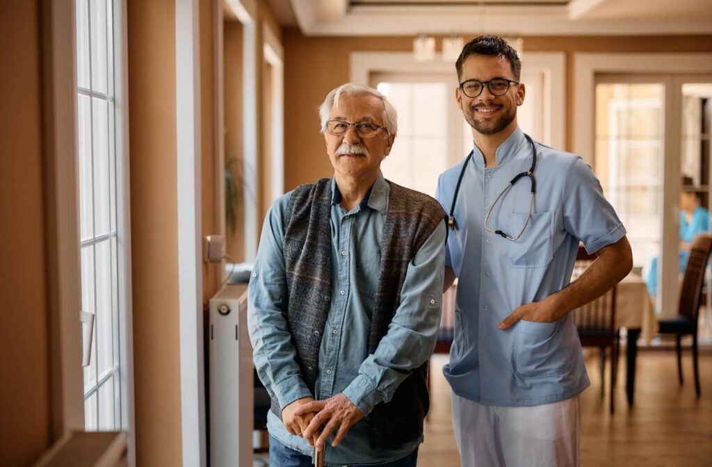 An older adult and a supportive caregiver stand beside one another in a beautiful hallway in an assisted living community