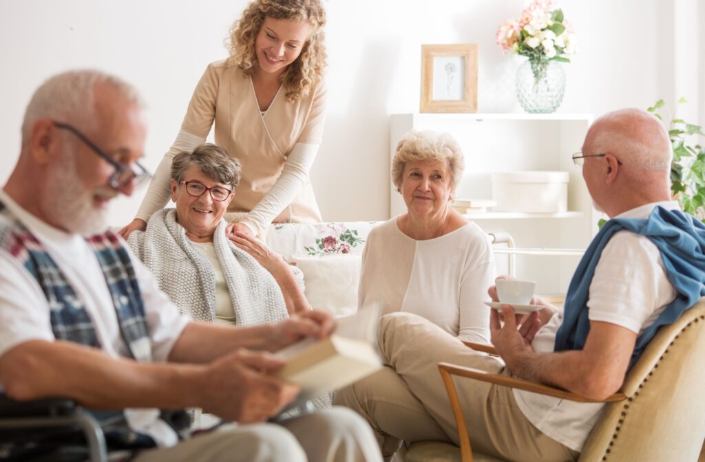 A group of 4 older adults sit reading and drinking coffee in assisted living while a caregiver checks in on them