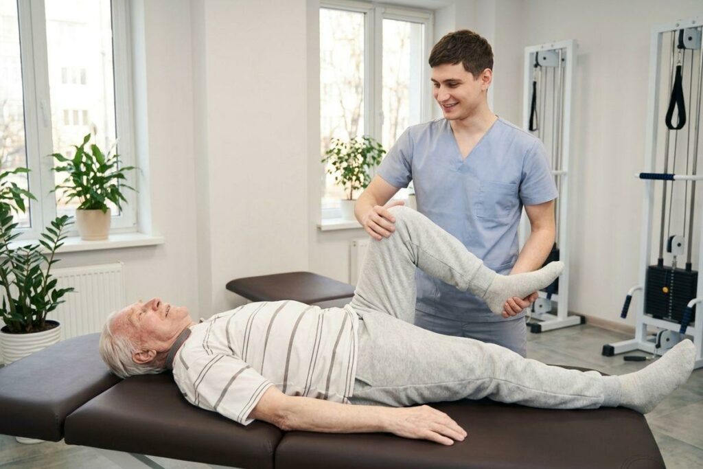 Physiotherapist assists an elderly man lying on a treatment table, lifting his leg for a stretch in a clinic.